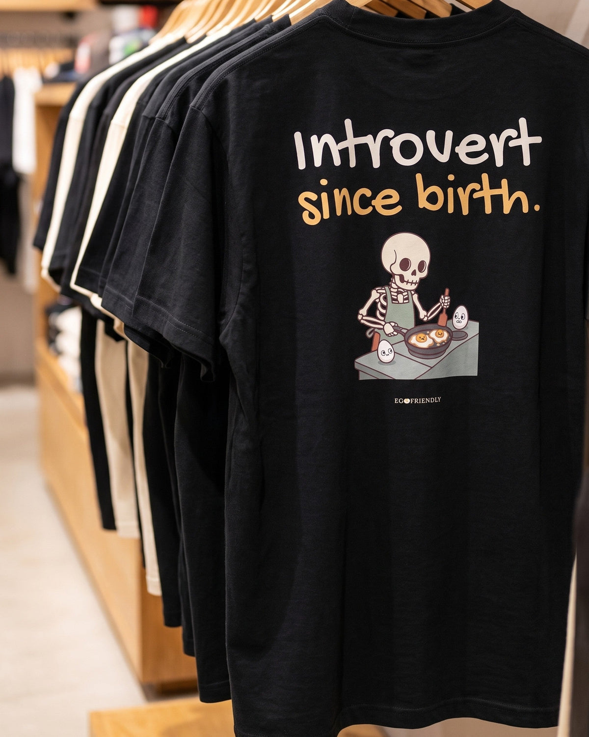 The Introvert Since Birth tee hanging on a rail in a clothing store, back print facing out: a skeleton frying two eggs while one egg stays whole.