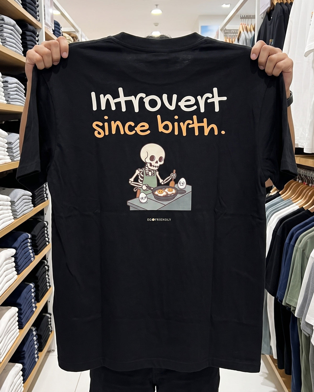 Someone holding the Introvert Since Birth tee open by the shoulders in a shop aisle, showing the full back print on black garment-dyed cotton.