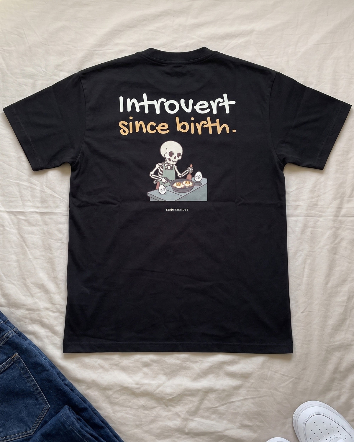 The Introvert Since Birth tee laid flat on a bed with jeans and white sneakers, back print facing up.