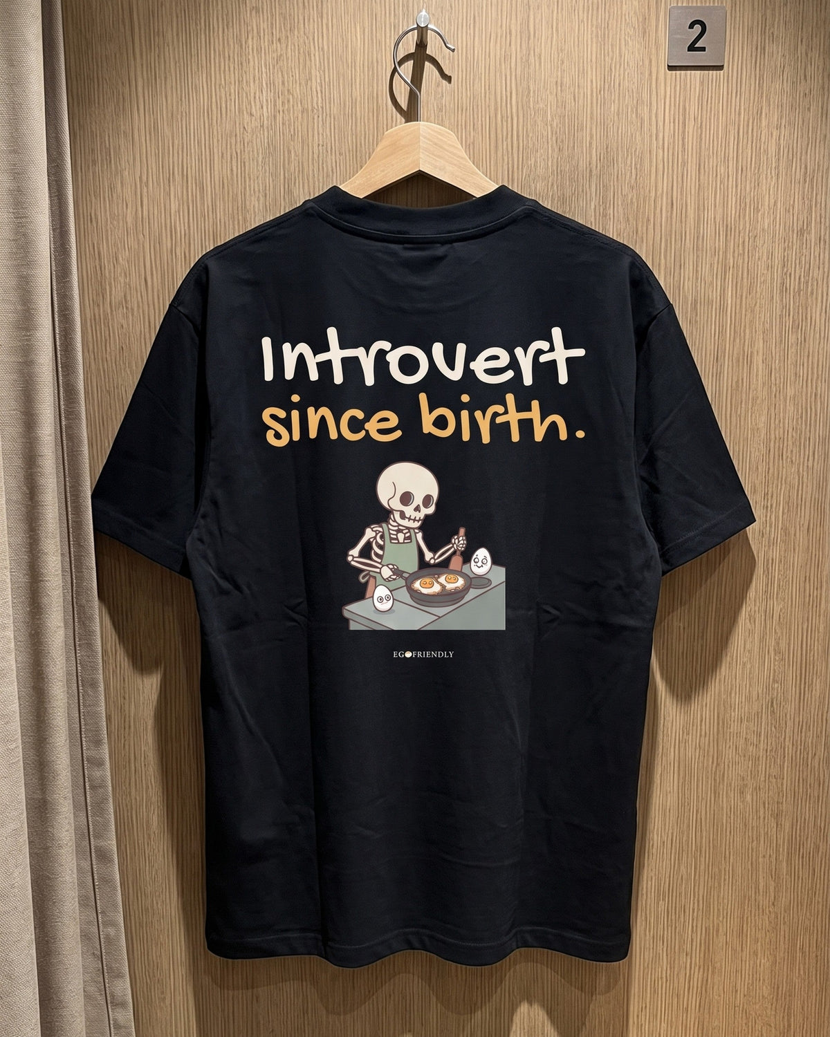 The Introvert Since Birth tee on a wooden hanger in a fitting room, back print facing the mirror.