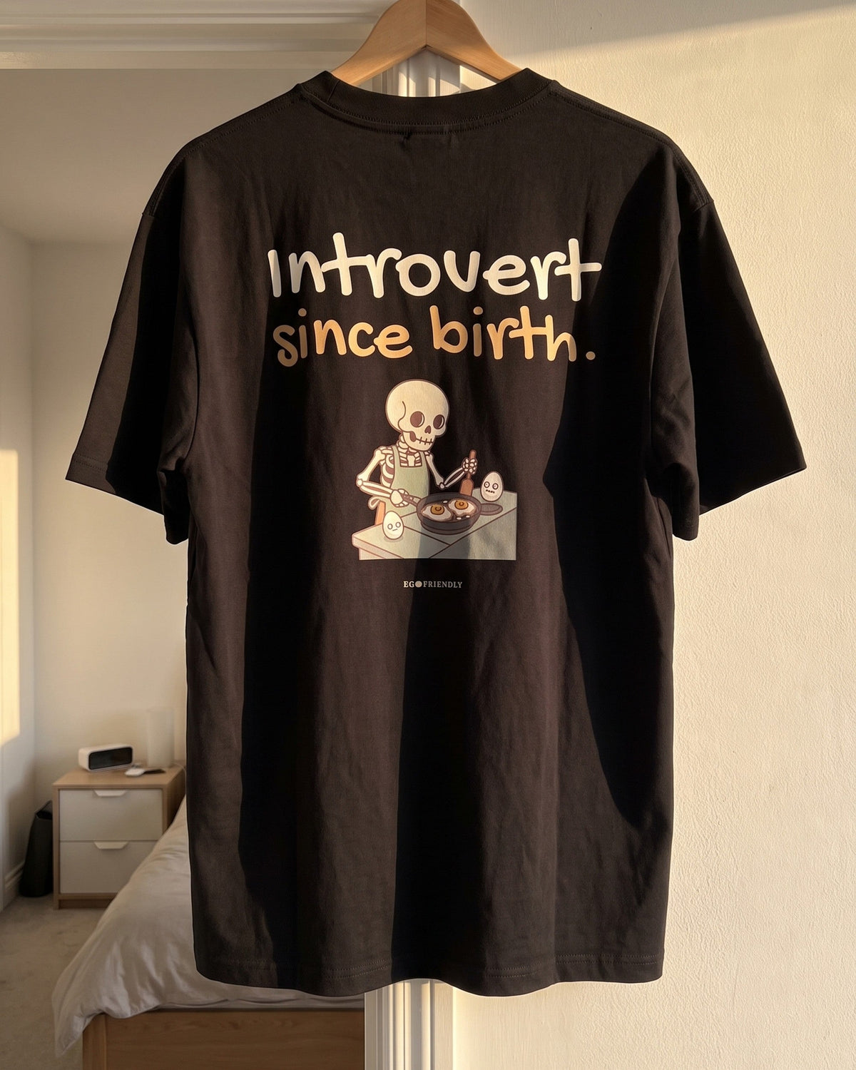 The Introvert Since Birth tee in pepper grey on a wooden hanger in a bedroom doorway, back print facing out.