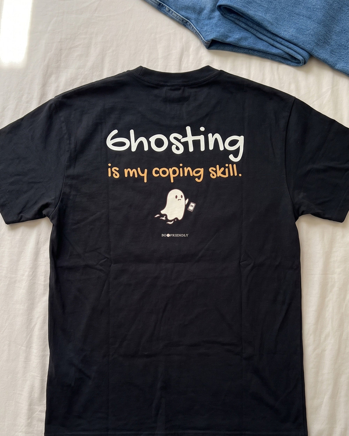The Ghosting Is My Coping Skill tee laid flat on a bed with jeans and white sneakers, back print facing up.