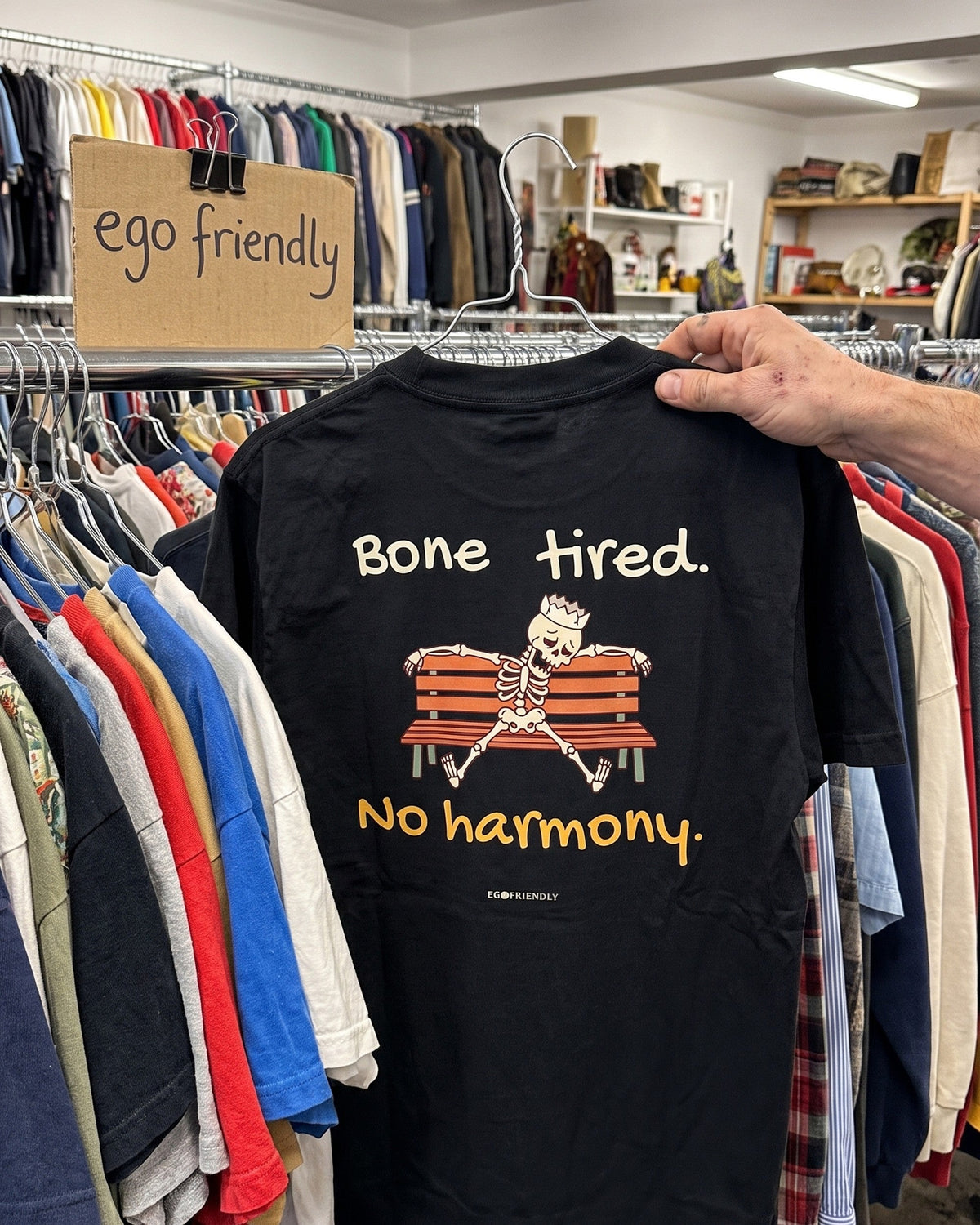 The Bone Tired No Harmony tee pulled forward from a thrift rail, back print facing out: an exhausted skeleton slumped on a park bench.