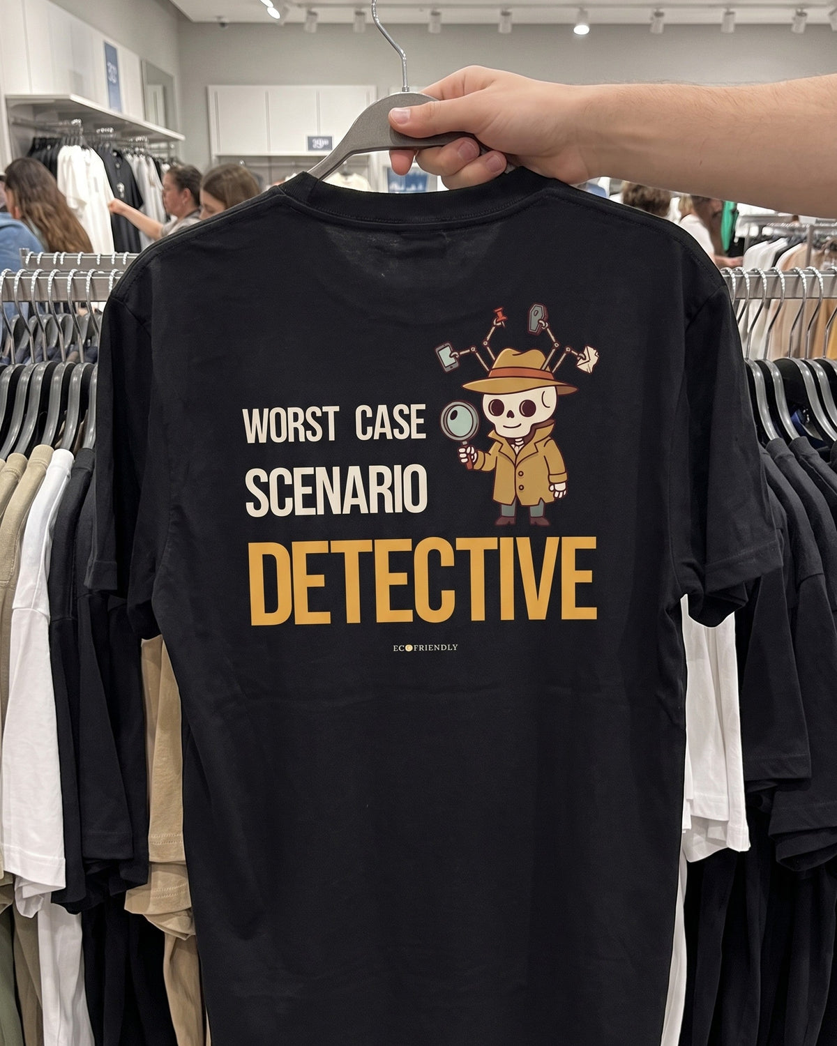 The Worst Case Scenario Detective tee on a hanger in a busy clothing store, showing the full back print on a black garment-dyed heavyweight tee.