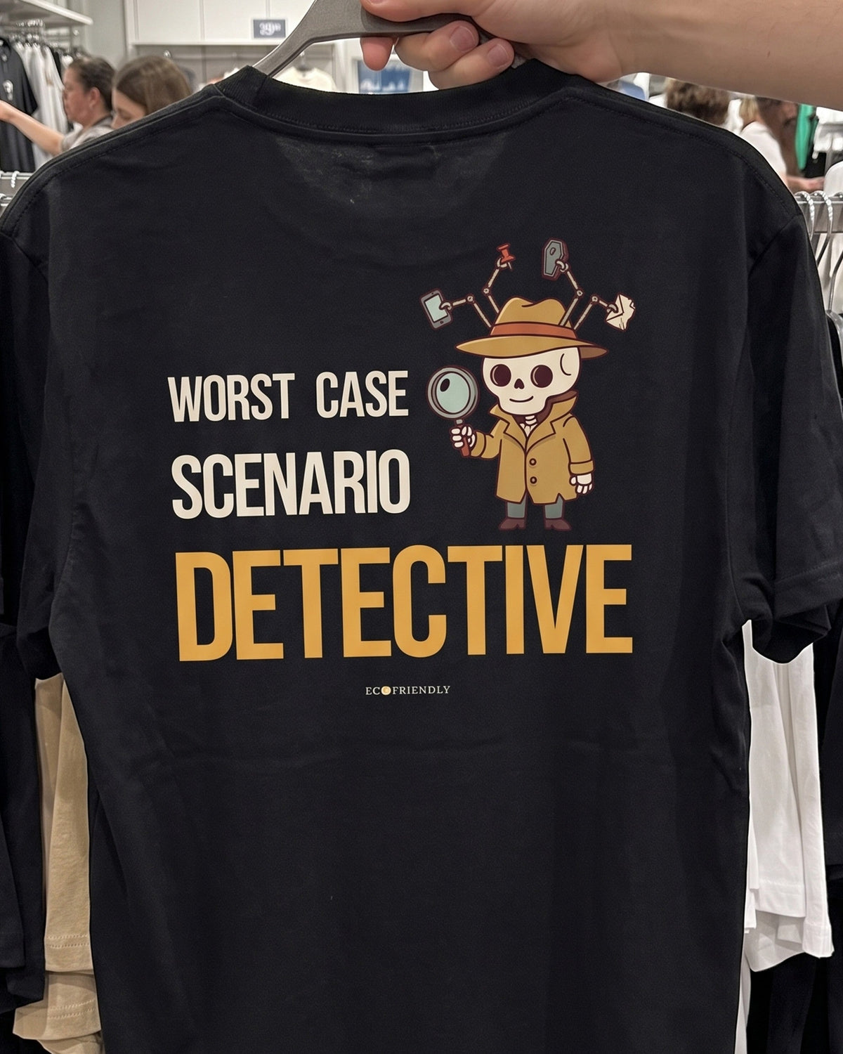 The Worst Case Scenario Detective tee held up on a hanger in a clothing store, back print facing out: a skeleton detective and his board of disasters that have not happened.