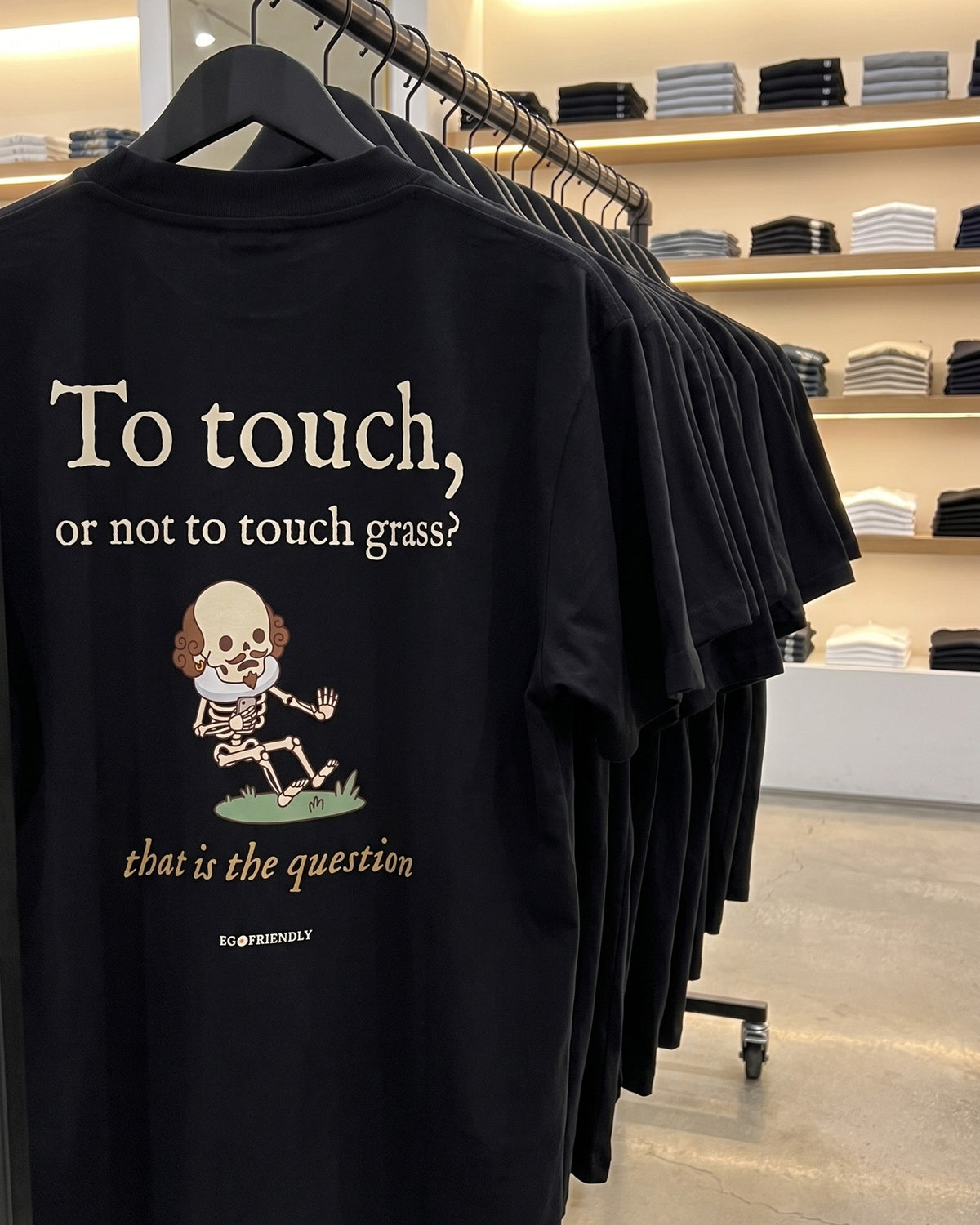 The To Touch Or Not To Touch Grass tee on a rail in a clothing store, back print facing out: Shakespeare as a skeleton recoiling from a patch of grass.