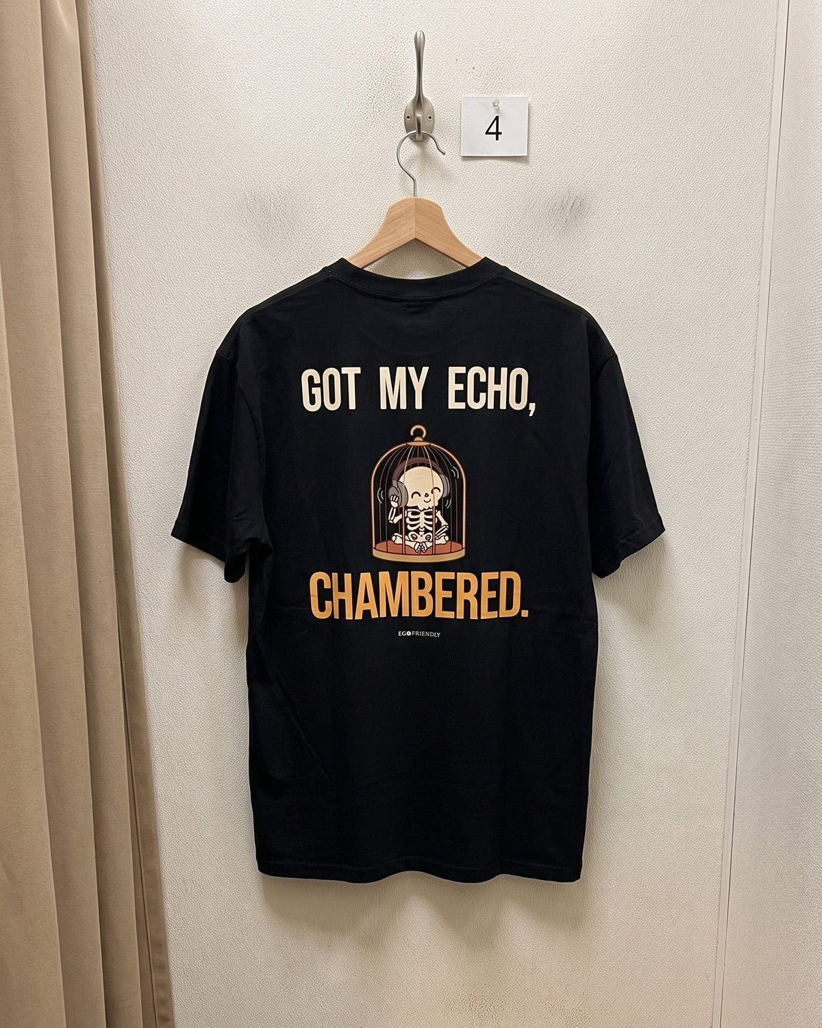 The Got My Echo Chambered tee on a wooden hanger against a plain wall, back print facing out: a skeleton in a birdcage wearing headphones.