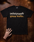 A black short-sleeve graphic T-shirt laid flat on a dark wooden surface, with a hand resting near the lower left hem. The shirt has the phrase 'Introvert since birth.