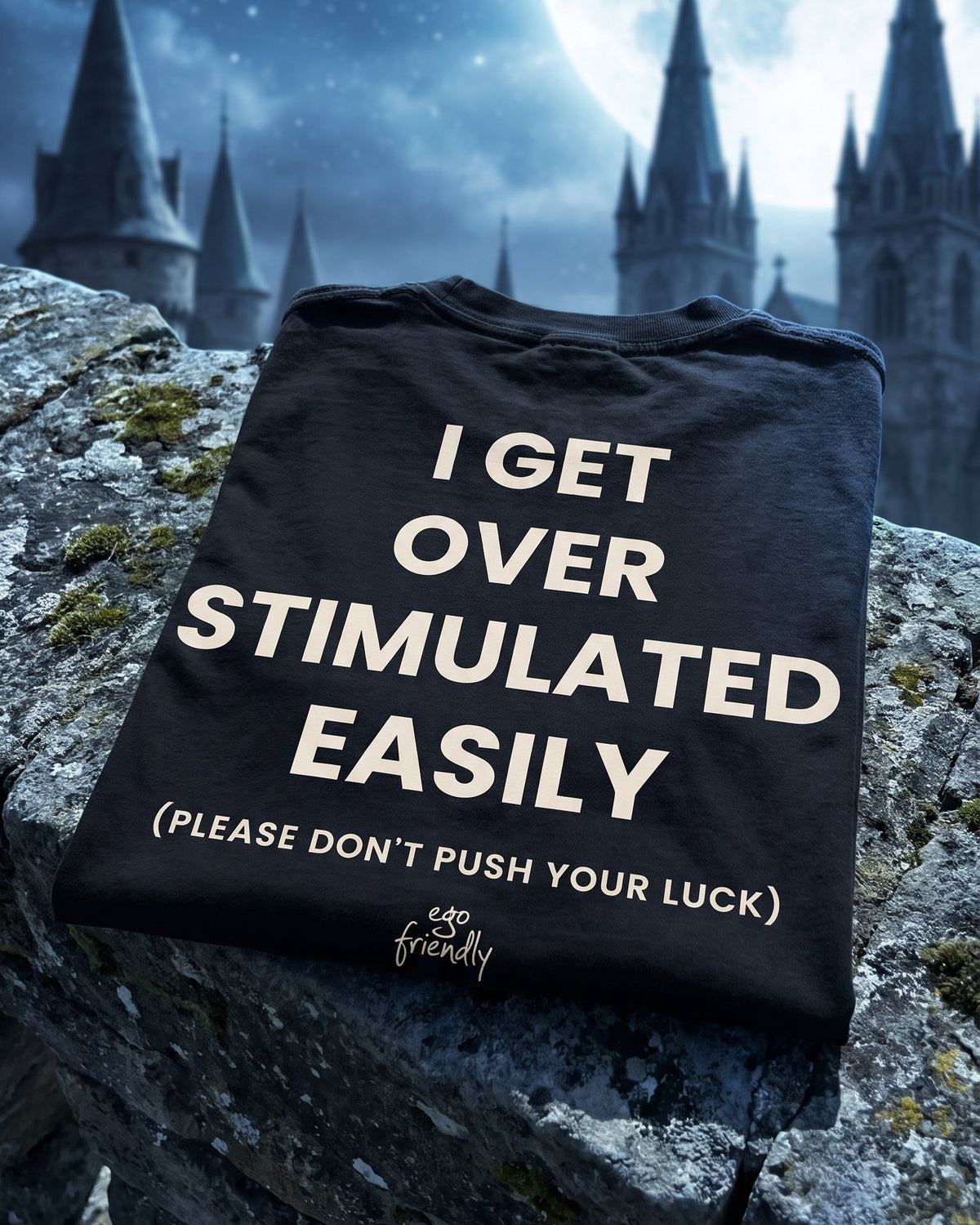 A black graphic t-shirt with cream text reading 'I GET OVER STIMULATED EASILY (PLEASE DON'T PUSH YOUR LUCK)' and a small 'ego friendly' logo, displayed on a stone surface with a dark castle and moonlit sky in the background.