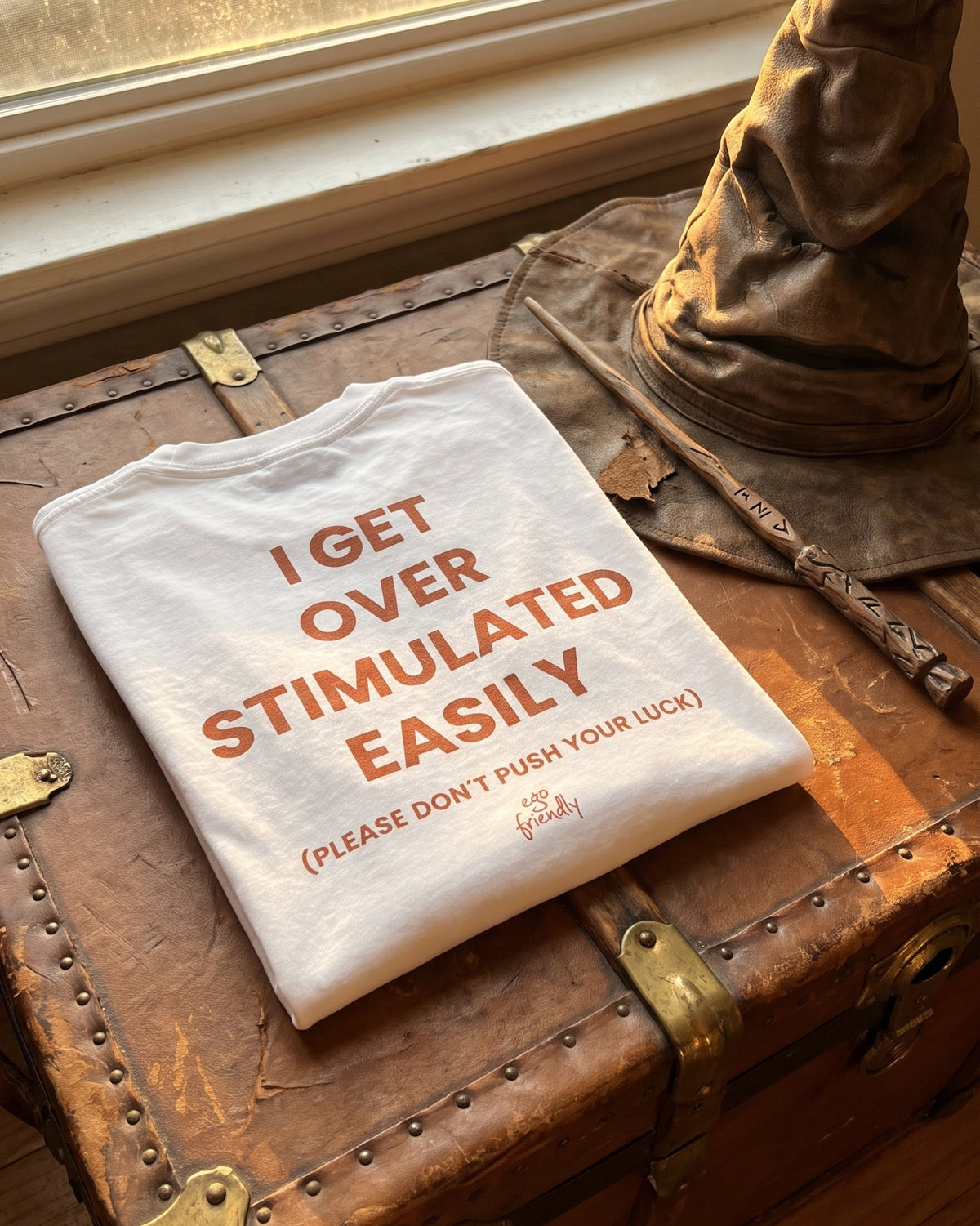 A folded white graphic T-shirt with orange text reading 'I GET OVER STIMULATED EASILY (PLEASE DON'T PUSH YOUR LUCK)' is displayed on a rustic brown leather trunk beside a brown hat, near a window.