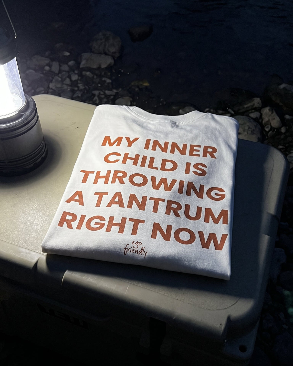 A white t-shirt with orange text reading 'MY INNER CHILD IS THROWING A TANTRUM RIGHT NOW' is laid on top of a cooler outdoors near water, with a lantern visible on the left.