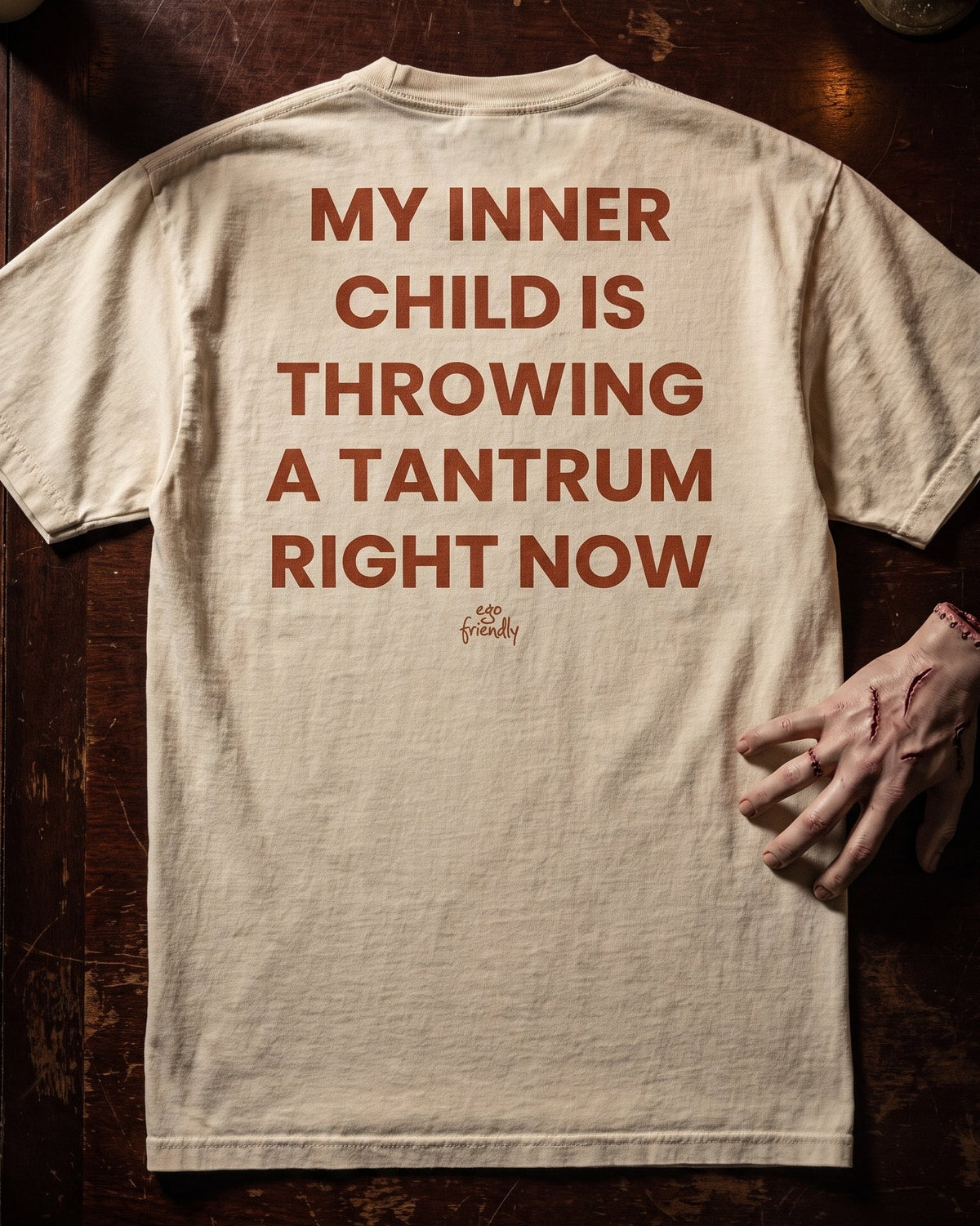 A cream-colored t-shirt with bold red text on the back reading 'MY INNER CHILD IS THROWING A TANTRUM RIGHT NOW', laid flat on a dark wooden table with a hand resting on the right side.