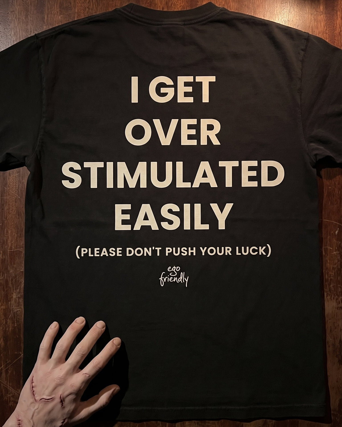A black short-sleeve T-shirt laid flat on a dark wood surface, showing the back with large cream text that reads 'I GET OVER STIMULATED EASILY (PLEASE DON'T PUSH YOUR LUCK)' and a small 'so friendly' logo below. A hand is placed at the lower left of the shirt.