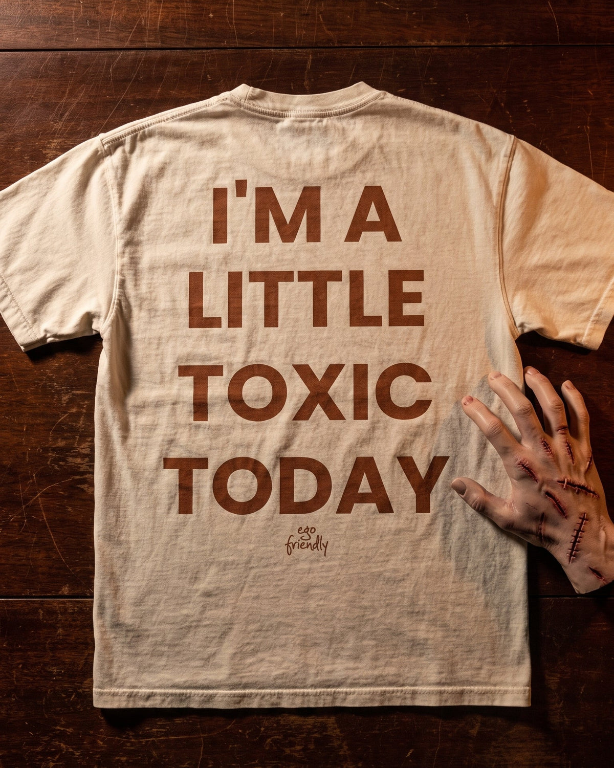 A beige crewneck t-shirt with the phrase 'I'M A LITTLE TOXIC TODAY' printed in brown block letters on the back, laid flat on a dark wood background next to a severed hand prop.