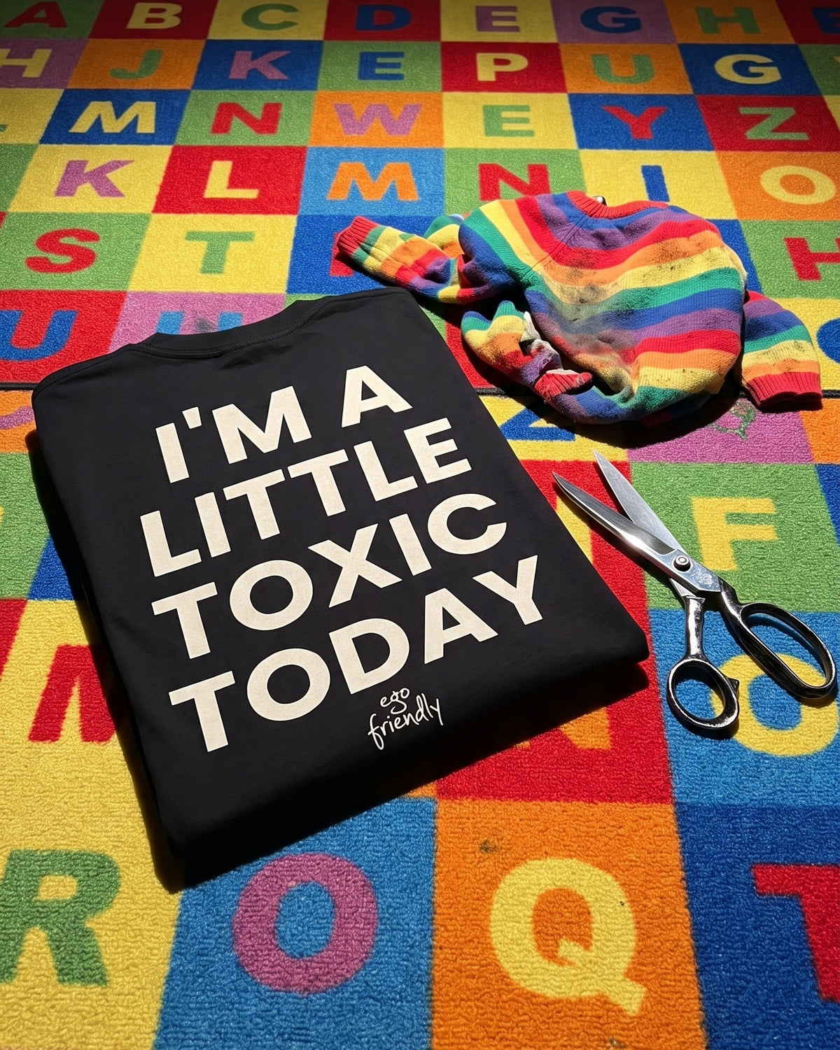 A folded black graphic T-shirt with the text 'I'M A LITTLE TOXIC TODAY' in large white letters, placed on a colorful alphabet rug alongside a rainbow-striped fabric item and metal scissors.