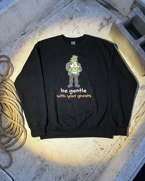 A black crewneck sweatshirt with a St. Patrick's Day graphic of a green-clad character and the text 'be gentle with your ghosts' in orange and white, laid flat on a concrete boat floor with a rope nearby.