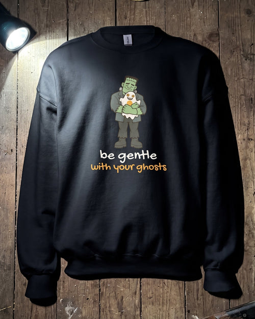 A black crewneck sweatshirt featuring a graphic of a green zombie-like ghost with the text 'be gentle with your ghosts' in white and orange, displayed on a rustic wooden background with a Jason Voorhees mask and a knife as props.