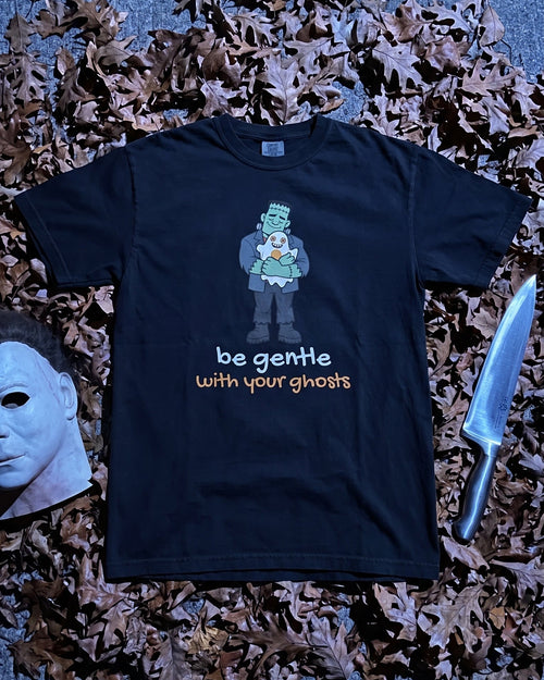 A black graphic t-shirt featuring a Frankenstein character and the text 'be gentle with your ghosts' in white and orange, styled in a flat lay on a gray concrete surface surrounded by brown autumn leaves, a Michael Myers mask, and a knife.