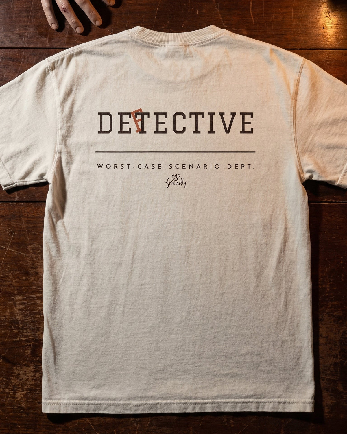 A light beige crewneck t-shirt laid flat on a dark wooden surface, showing the back with a graphic that reads 'DETECTIVE' and 'WORST-CASE SCENARIO DEPT.' with a small 'the fantasty' logo.