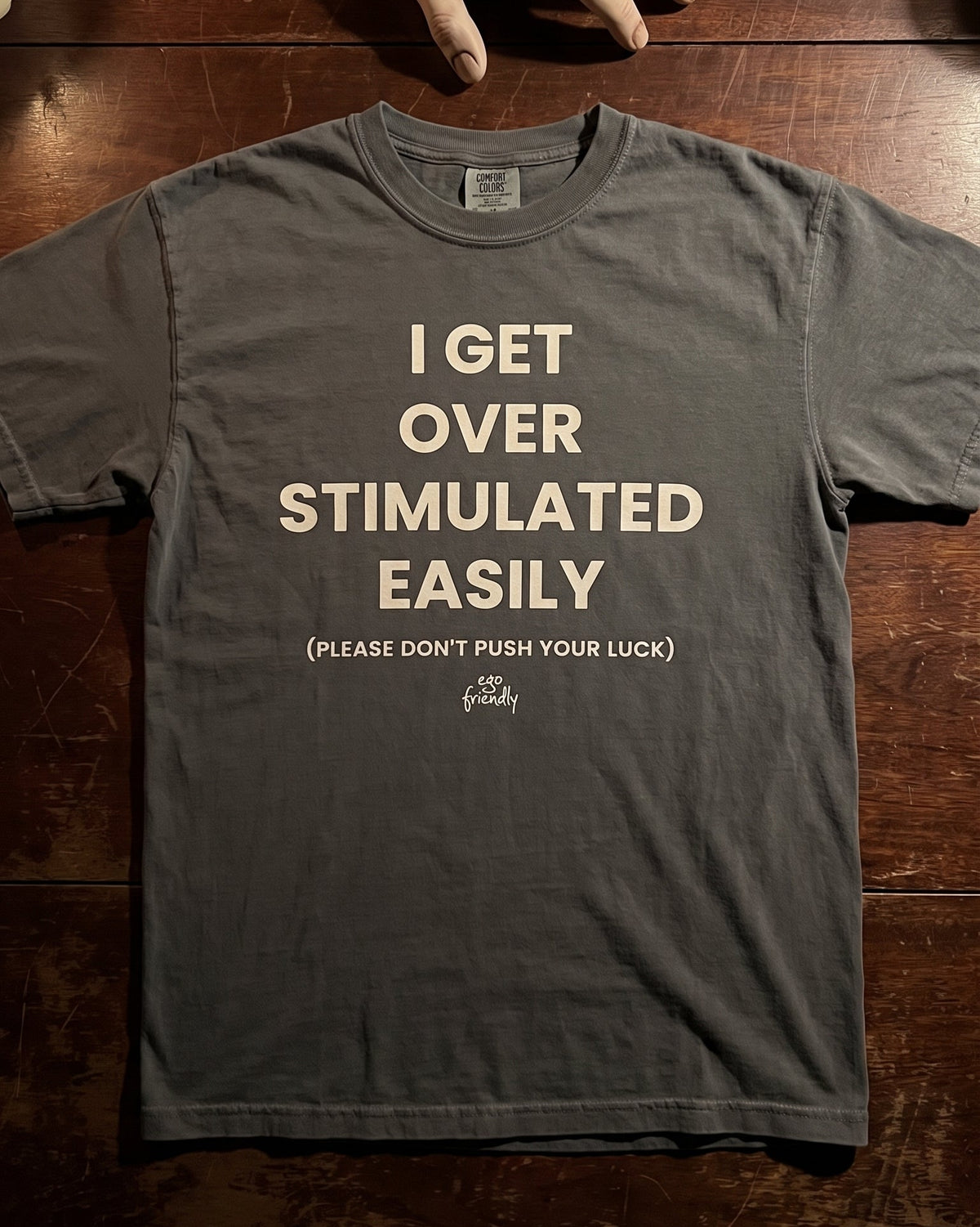 A dark gray graphic t-shirt laid flat on a dark wooden surface, featuring a severed hand prop above it. The t-shirt has cream-colored text that reads 'I GET OVER STIMULATED EASILY (PLEASE DON'T PUSH YOUR LUCK)'.