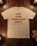 A beige graphic t-shirt with brown text reading 'I GET OVER STIMULATED EASILY (PLEASE DON'T PUSH YOUR LUCK)' is laid flat on a dark wooden surface with candles and a hand visible at the edge.