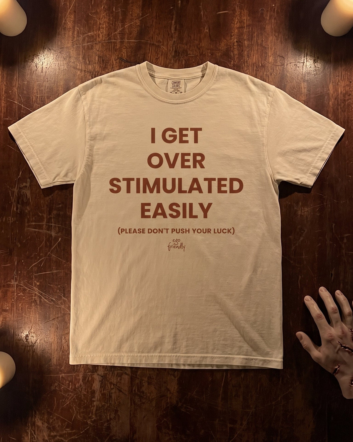 A beige graphic t-shirt with brown text reading 'I GET OVER STIMULATED EASILY (PLEASE DON'T PUSH YOUR LUCK)' is laid flat on a dark wooden surface with candles and a hand visible at the edge.