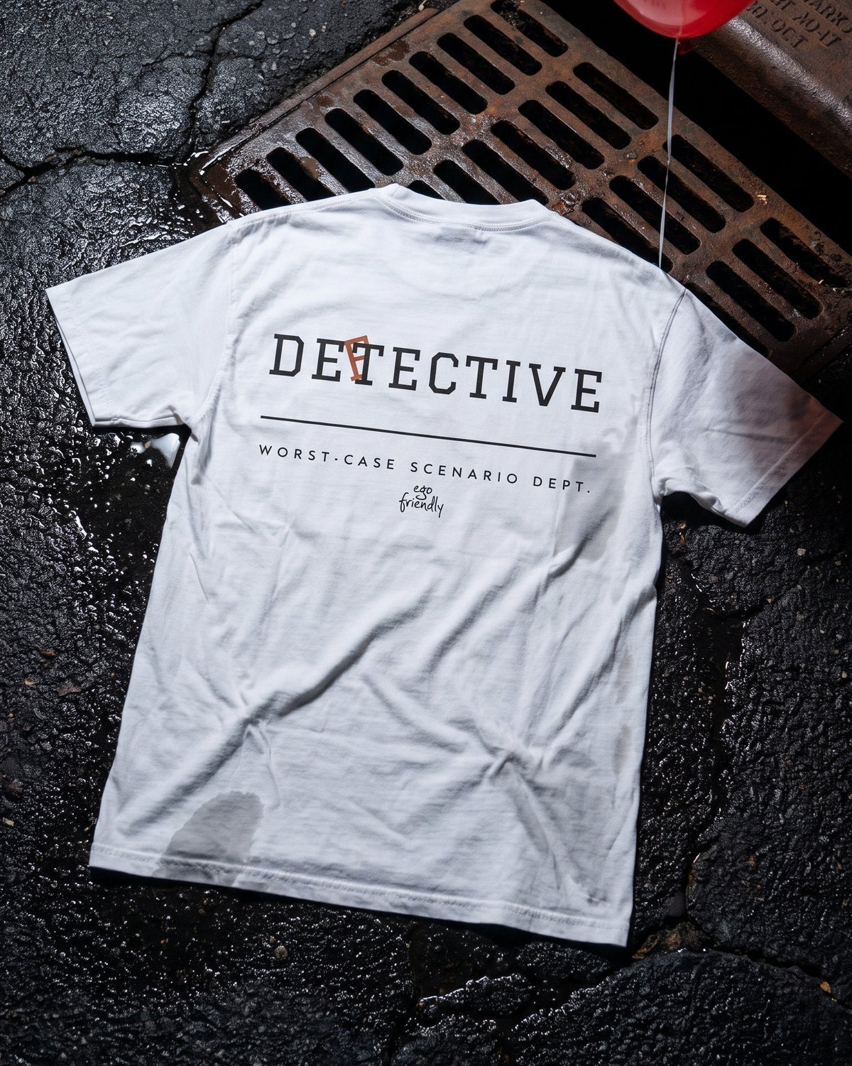 A white graphic t-shirt laid flat on wet asphalt next to a metal drain grate. The back of the shirt features black text reading 'DETECTIVE' with a small orange accent, and 'WORST-CASE SCENARIO DEPT.