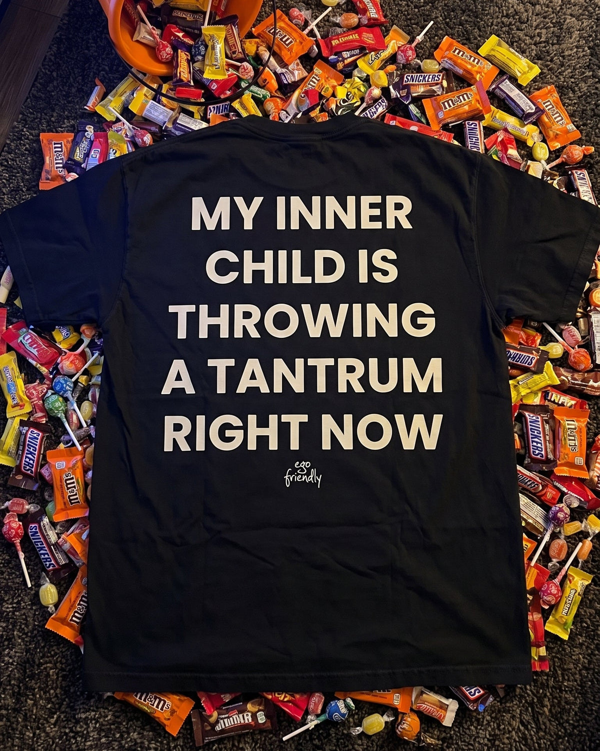 A black short-sleeve T-shirt laid flat on a carpeted floor, surrounded by an abundant pile of assorted candy. The back of the shirt features large, bold white text that reads 'MY INNER CHILD IS THROWING A TANTRUM RIGHT NOW' with a small cursive brand signature below.