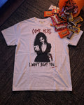 A white graphic t-shirt featuring a gothic-style illustration of a woman with the text 'COME HERE I WON'T BEAT YOU' laid flat on a dark carpet. An orange pumpkin bucket filled with candy sits to the upper right.