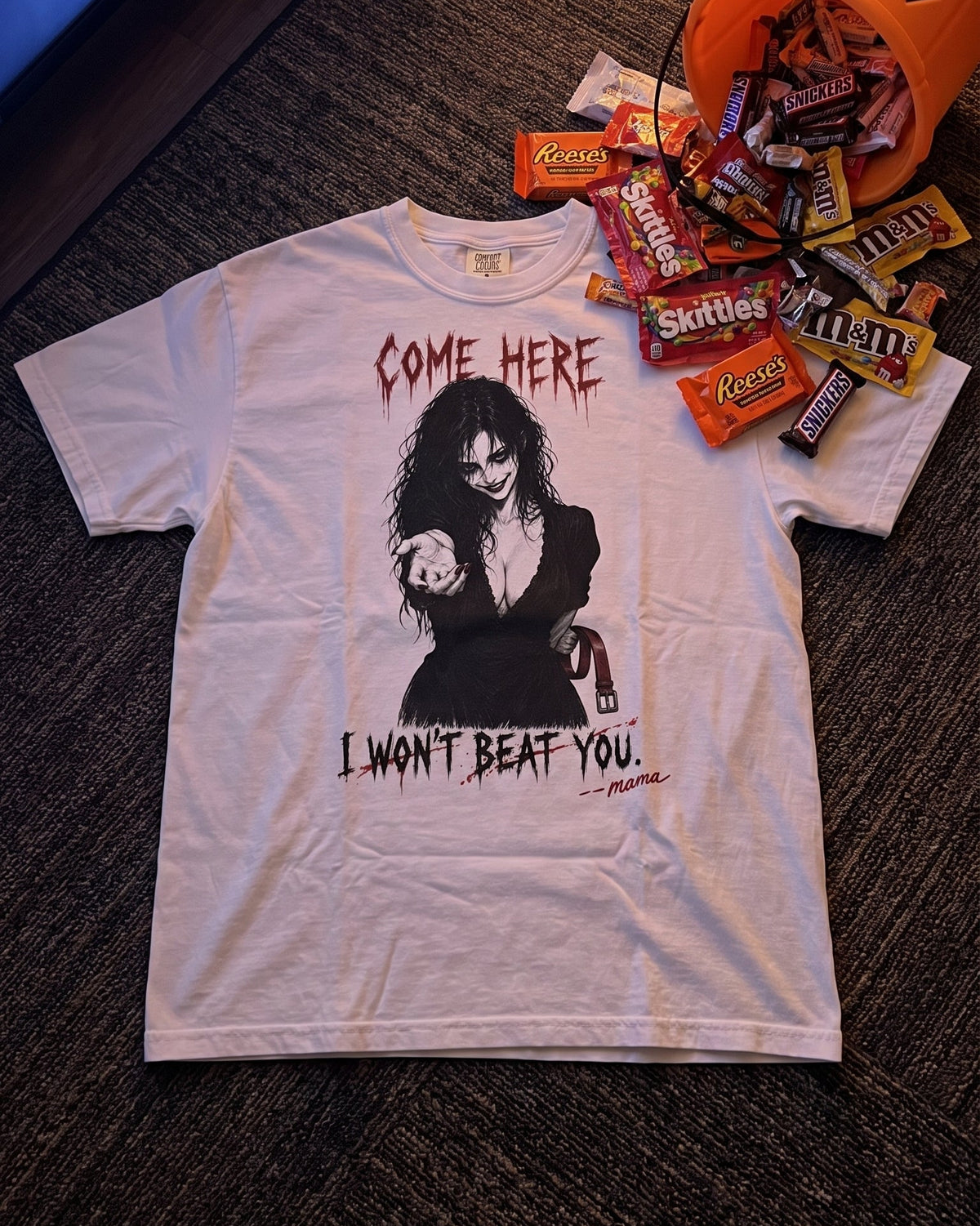 A white graphic t-shirt featuring a gothic-style illustration of a woman with the text 'COME HERE I WON'T BEAT YOU' laid flat on a dark carpet. An orange pumpkin bucket filled with candy sits to the upper right.