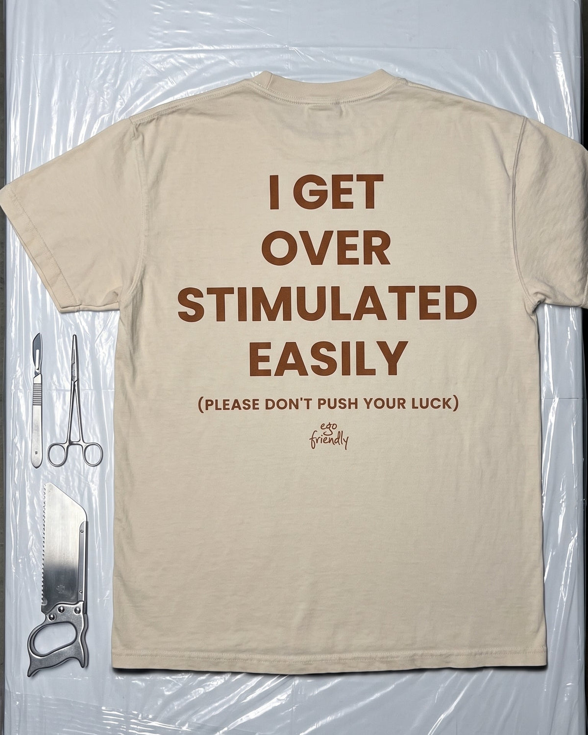 A beige graphic t-shirt laid flat on a clear plastic sheet, featuring the humorous phrase 'I GET OVER STIMULATED EASILY (PLEASE DON'T PUSH YOUR LUCK)' in brown text on the back.