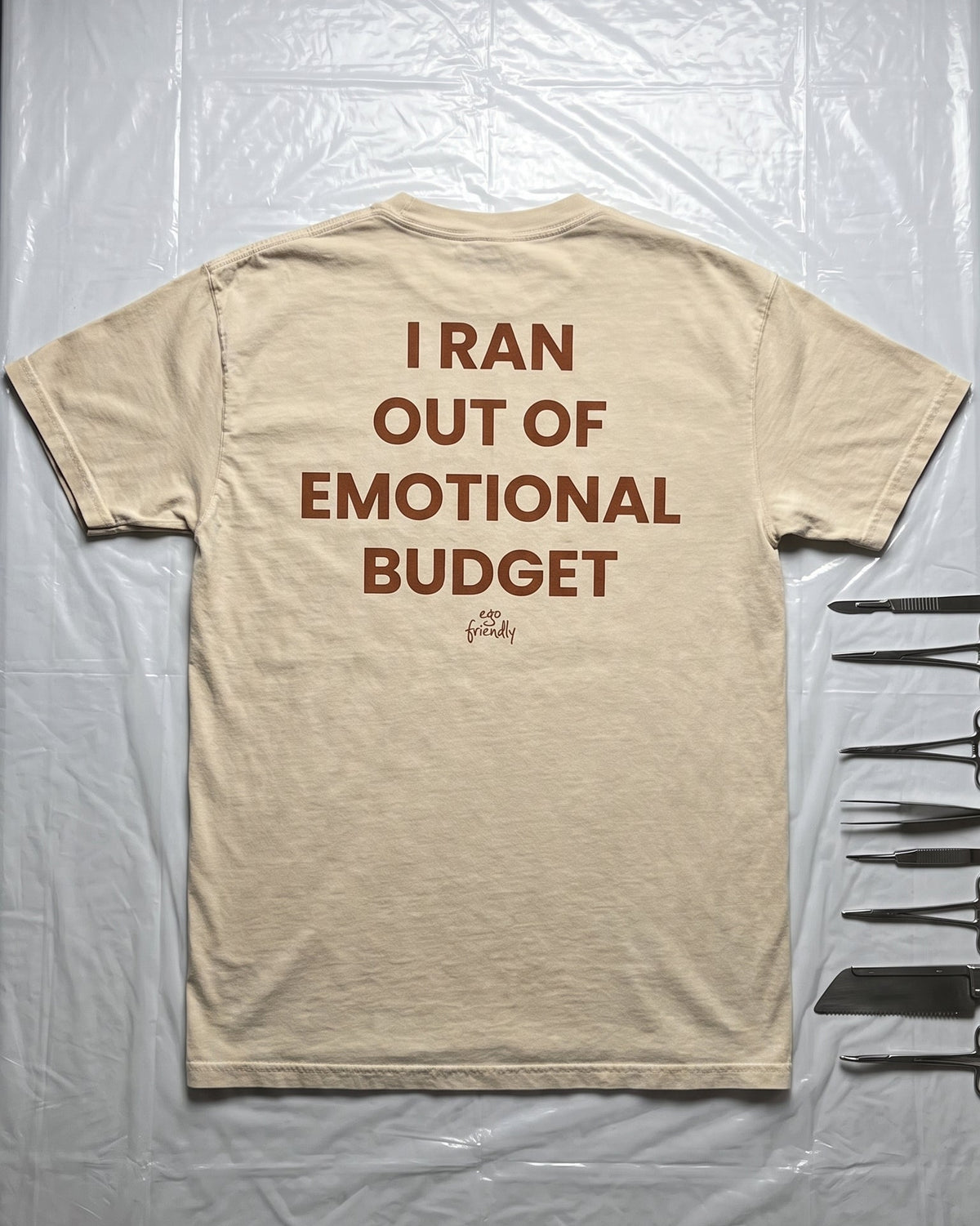 A beige crewneck t-shirt laid flat on a wrinkled white plastic sheet, featuring the humorous text 'I RAN OUT OF EMOTIONAL BUDGET' printed in brown on the back.