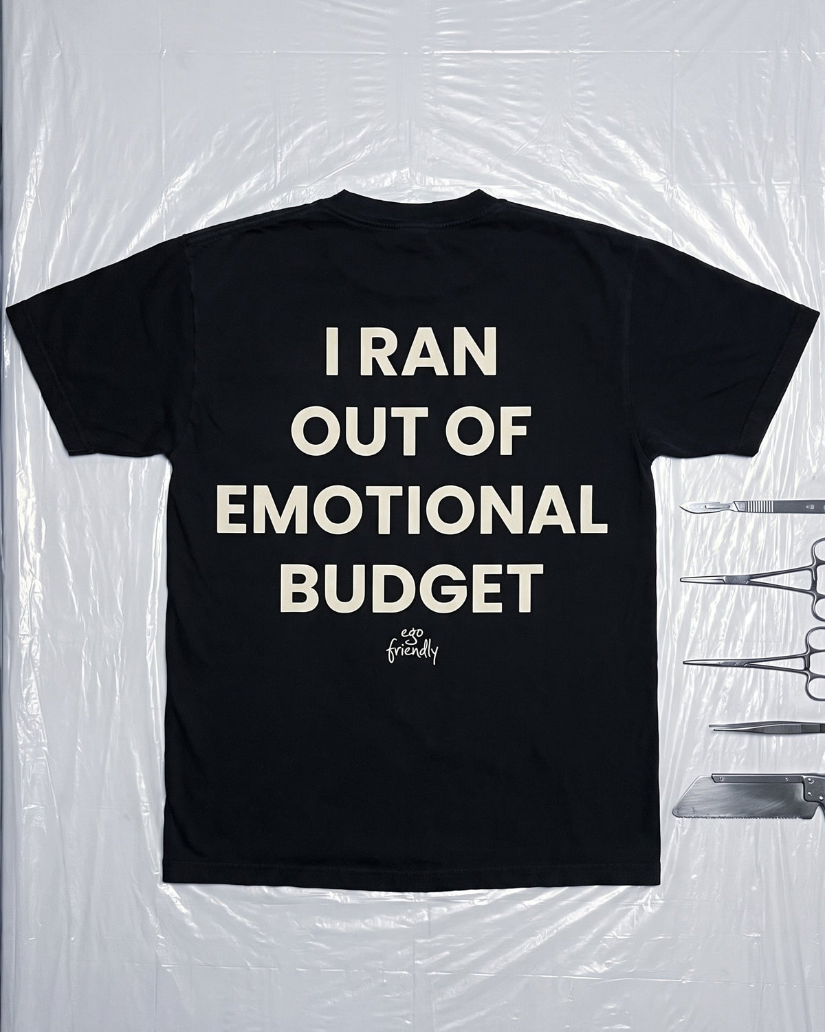 A black short-sleeve t-shirt with the phrase 'I RAN OUT OF EMOTIONAL BUDGET' printed in bold cream lettering, laid flat on a white surface wrapped in clear plastic.