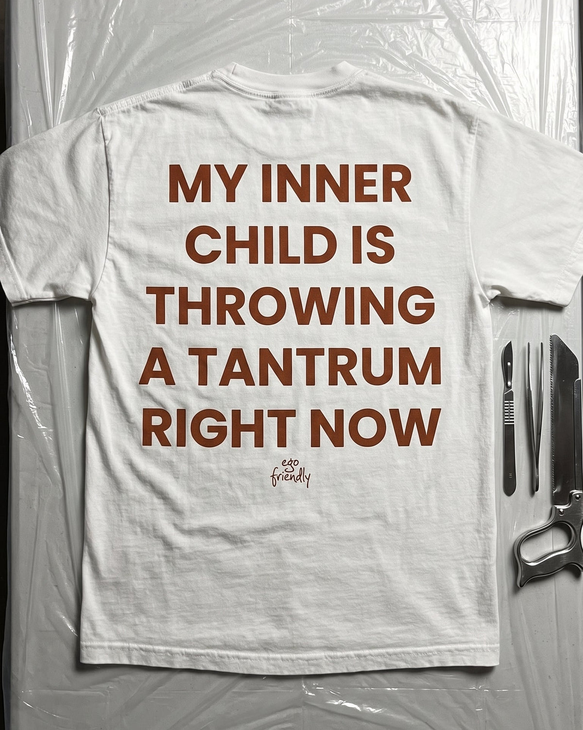 A white graphic t-shirt with bold brown text reading 'MY INNER CHILD IS THROWING A TANTRUM RIGHT NOW' and a small 'The Happy.