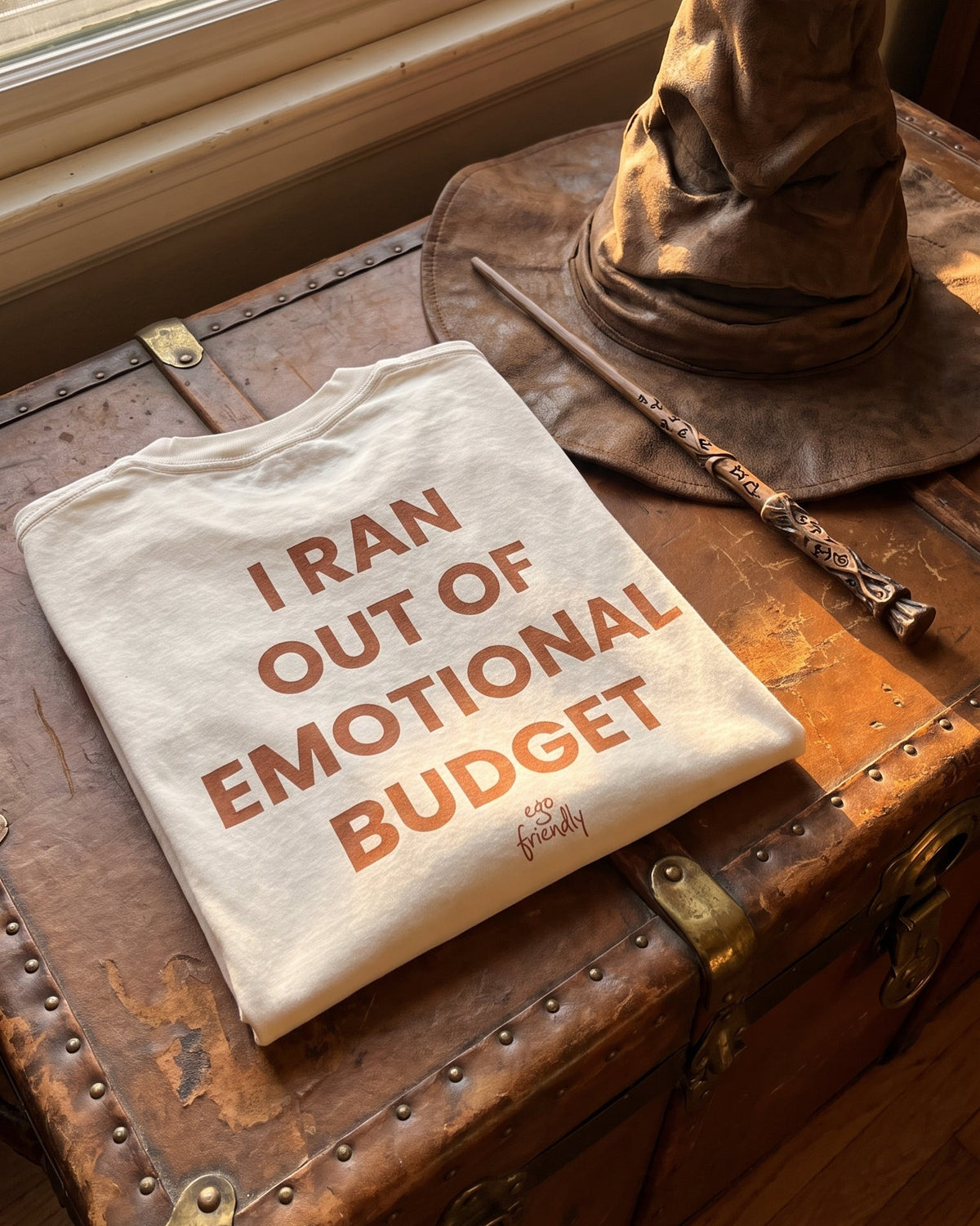A folded cream-colored t-shirt with orange text reading 'I RAN OUT OF EMOTIONAL BUDGET' is displayed on a rustic brown leather trunk next to a wizard hat and wand in front of a window.