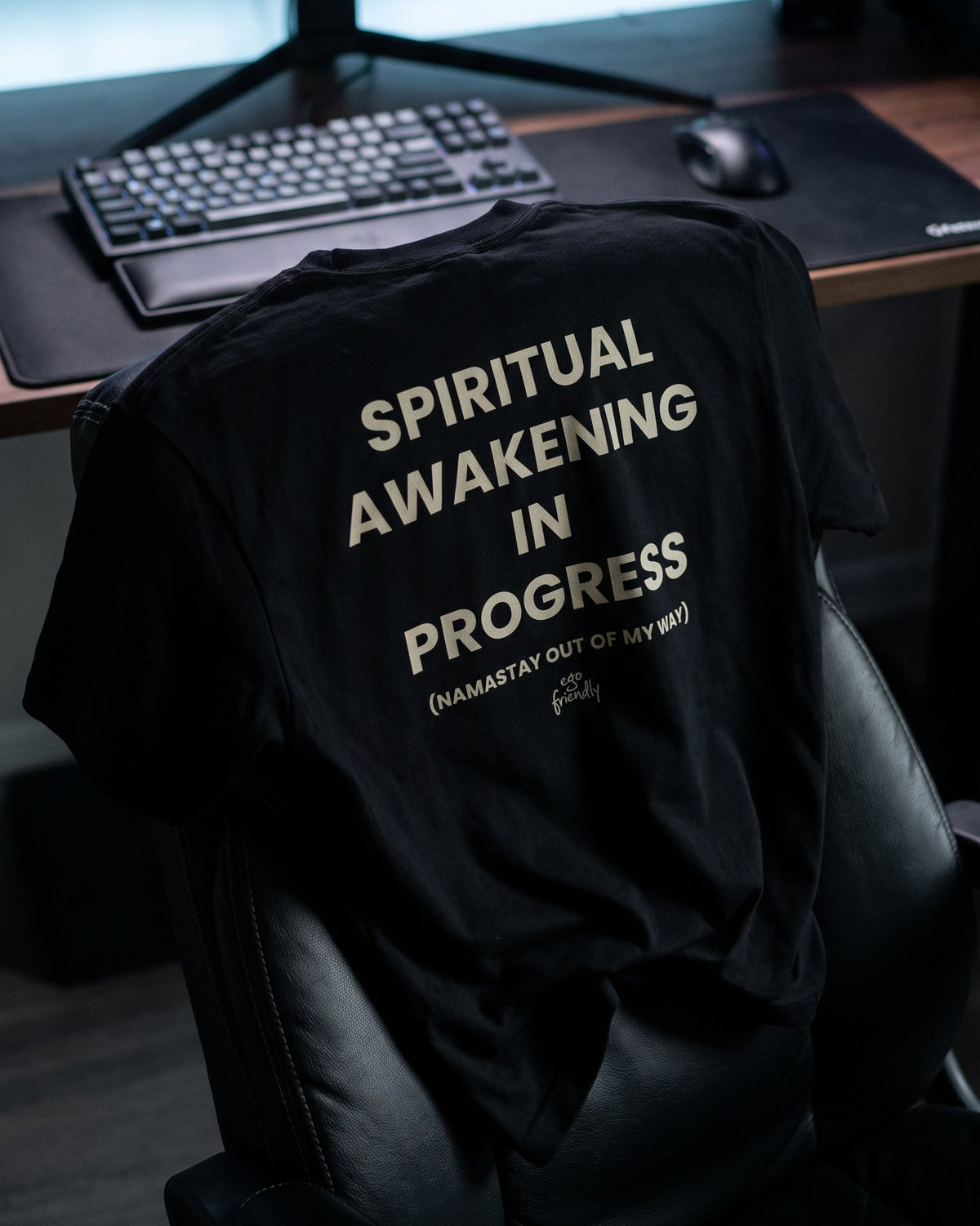 A black graphic t-shirt draped over a black gaming chair in front of a computer desk setup. The shirt features the text 'SPIRITUAL AWAKENING IN PROGRESS' and '(HAMSAY OUT OF MY HEAD)' printed in a light beige font.
