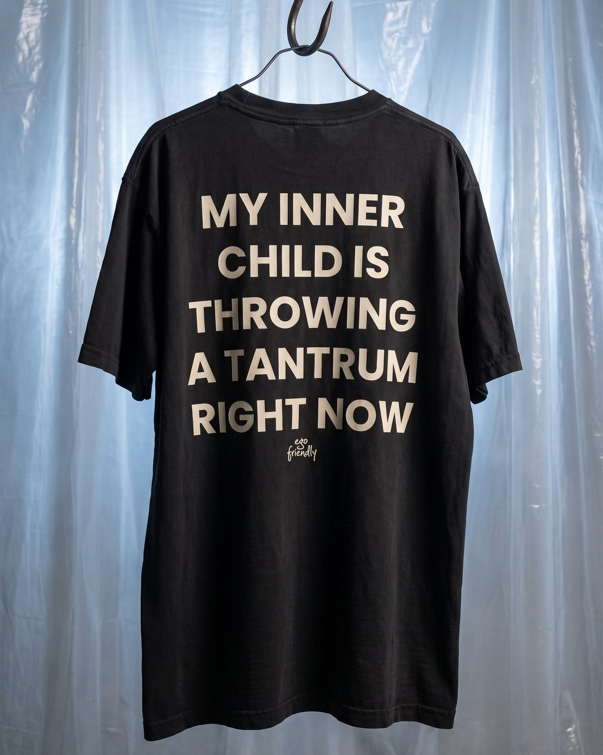 A black short-sleeve t-shirt hanging on a metal hanger against a backdrop of translucent blue plastic sheeting. The back of the shirt features a large, distressed cream-colored text graphic that reads 'MY INNER CHILD IS THROWING A TANTRUM RIGHT NOW'.