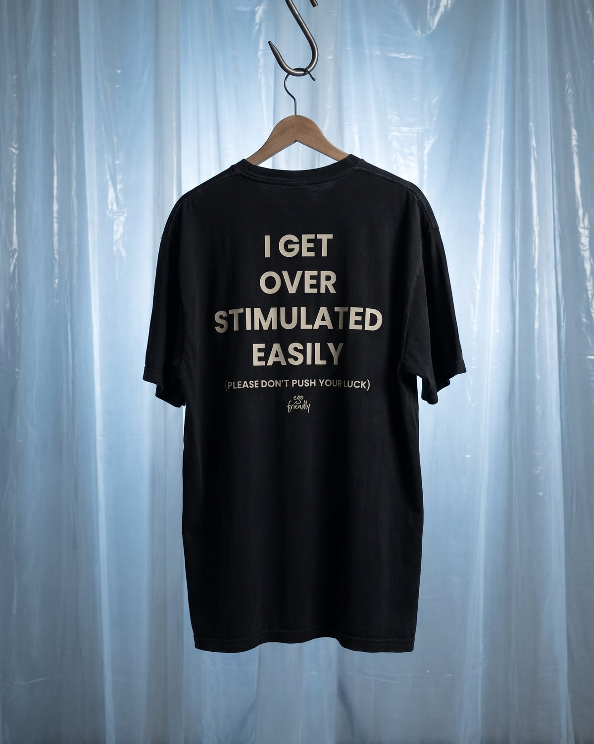 A black short-sleeve t-shirt hanging on a wooden hanger against a light blue translucent curtain backdrop. The back of the shirt features bold white text that reads 'I GET OVER STIMULATED EASILY' with smaller text below reading 'PLEASE DON'T PUSH YOUR LUCK' and a small script logo underneath.
