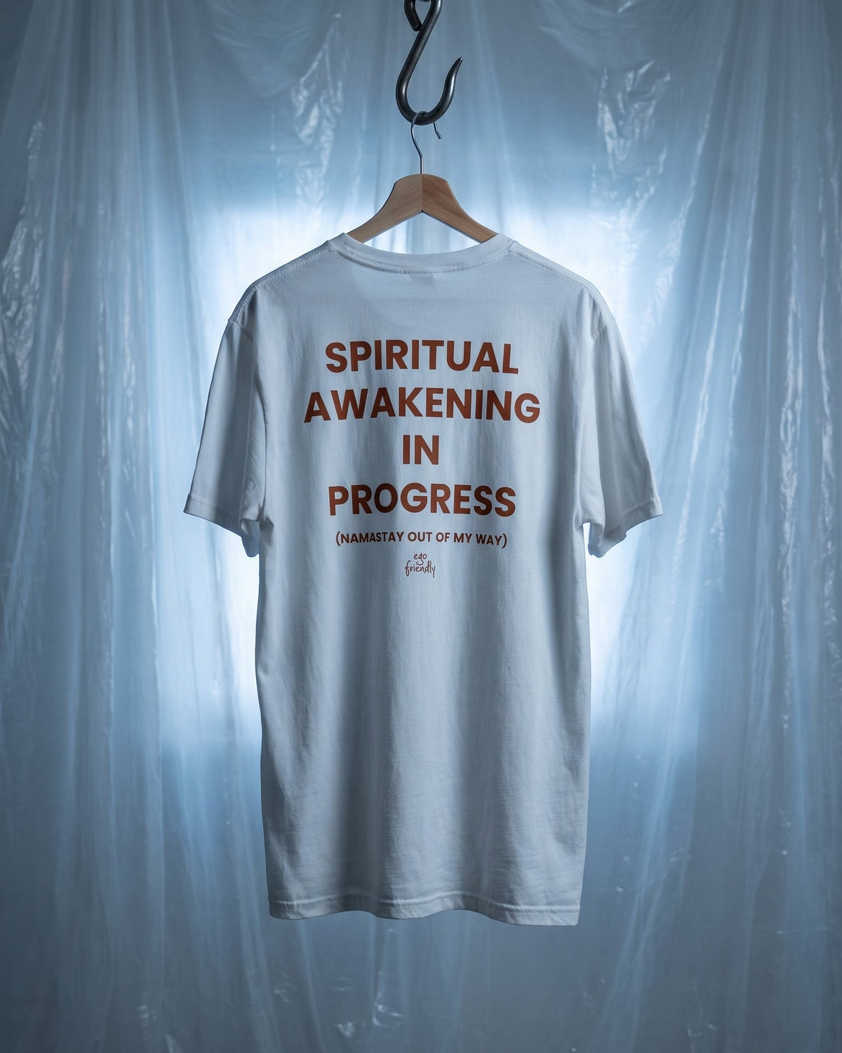 A white short-sleeve t-shirt hanging on a wooden hanger from a black hook, set against a backdrop of draped translucent plastic sheeting with cool blue lighting. The back of the shirt features the text 'SPIRITUAL AWAKENING IN PROGRESS' in a reddish-brown font, with smaller text below reading 'ENLIGHTEN OUT OF MY MIND' and a small signature.