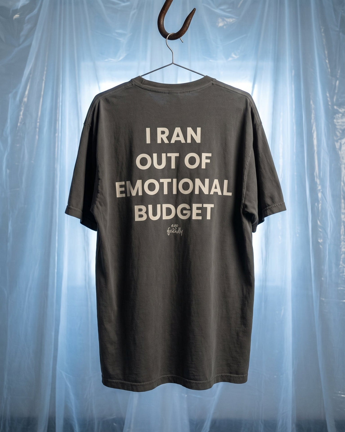 A dark gray t-shirt hanging from a rusty hook on a hanger against a backdrop of translucent blue plastic sheeting. The back of the shirt features the text 'I RAN OUT OF EMOTIONAL BUDGET' in a light beige font.