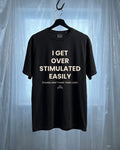 A black short-sleeve graphic t-shirt hanging on a black hanger from a metal hook, set against a backdrop of translucent plastic sheeting. The shirt features white text that reads 'I GET OVER STIMULATED EASILY (PLEASE DON'T PUSH YOUR LUCK)' with a small 'Grit' brand logo at the bottom.