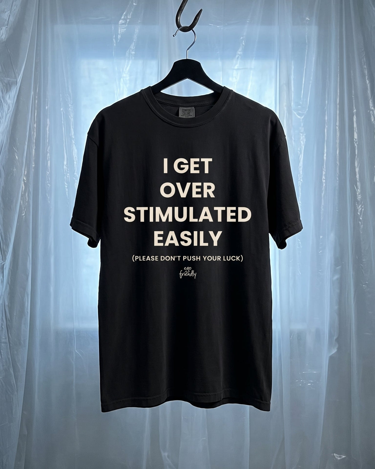 A black short-sleeve graphic t-shirt hanging on a black hanger from a metal hook, set against a backdrop of translucent plastic sheeting. The shirt features white text that reads 'I GET OVER STIMULATED EASILY (PLEASE DON'T PUSH YOUR LUCK)' with a small 'Grit' brand logo at the bottom.