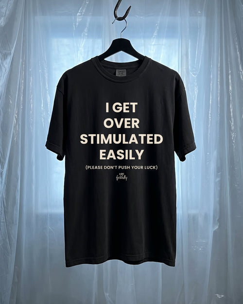 A black short-sleeve graphic t-shirt hanging on a black hanger from a metal hook, set against a backdrop of translucent plastic sheeting. The shirt features white text that reads 'I GET OVER STIMULATED EASILY (PLEASE DON'T PUSH YOUR LUCK)' with a small 'Grit' brand logo at the bottom.
