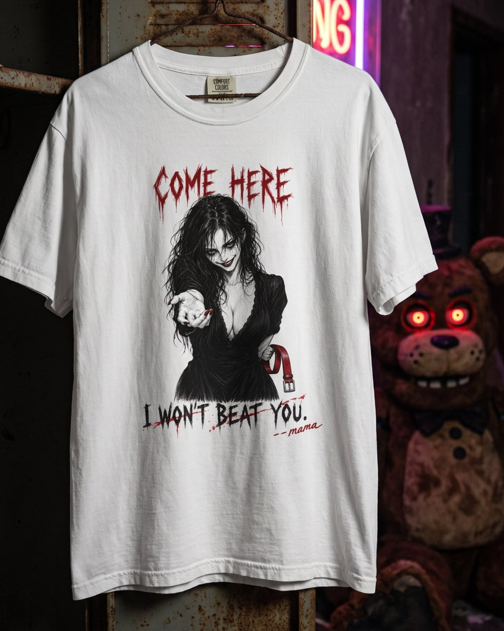 A white graphic t-shirt hanging on a rusty metal door in a dark, atmospheric room. The shirt features a black and red graphic of a woman holding a knife, with the text 'COME HERE' and 'I WON'T BEAT YOU.