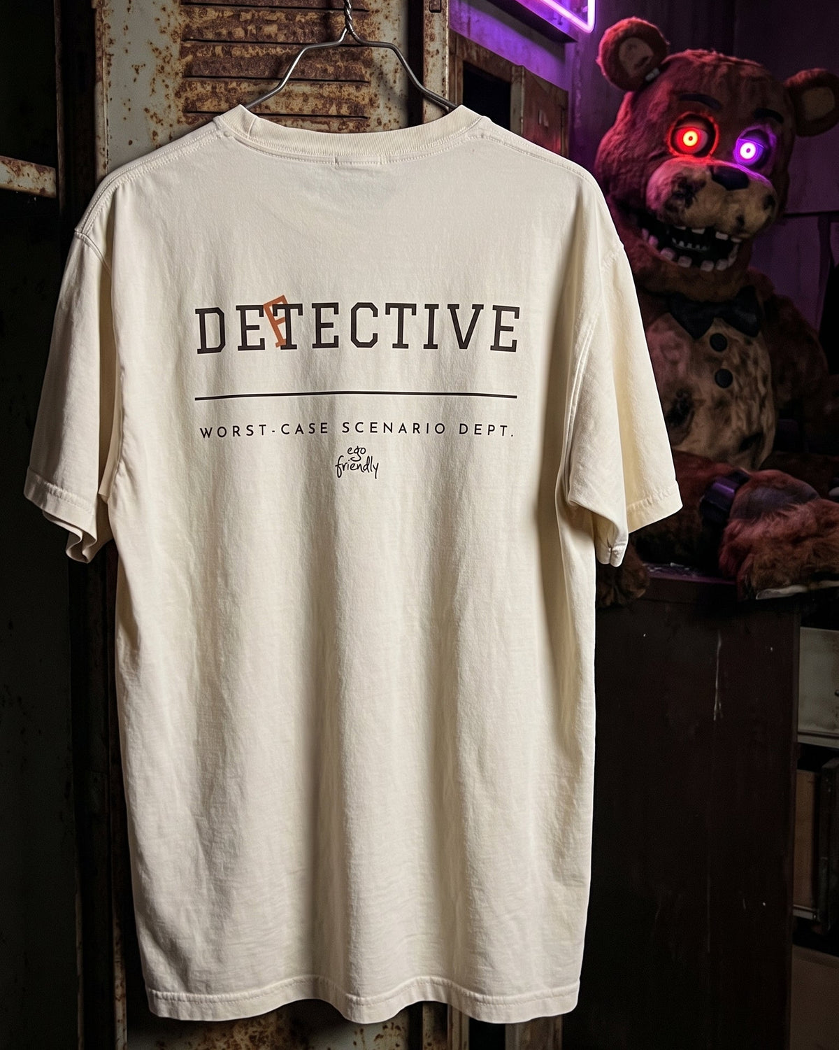 An off-white graphic t-shirt hanging on a metal hanger against a dark, rustic background with a Freddy Krueger prop. The shirt features the word 'DETECTIVE' in black with a small orange accent, and 'WORST-CASE SCENARIO DEPT.