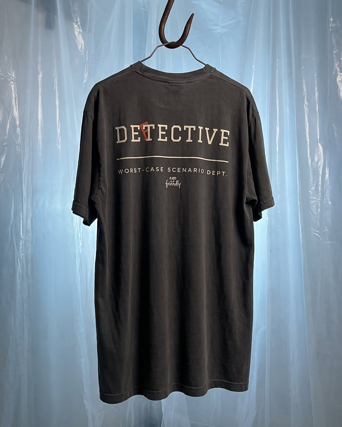A dark brown short-sleeve t-shirt hanging from a metal hook against a backdrop of translucent plastic sheeting. The shirt features 'DETECTIVE' printed in beige and red across the chest, with 'Worst Case Scenario Dept.