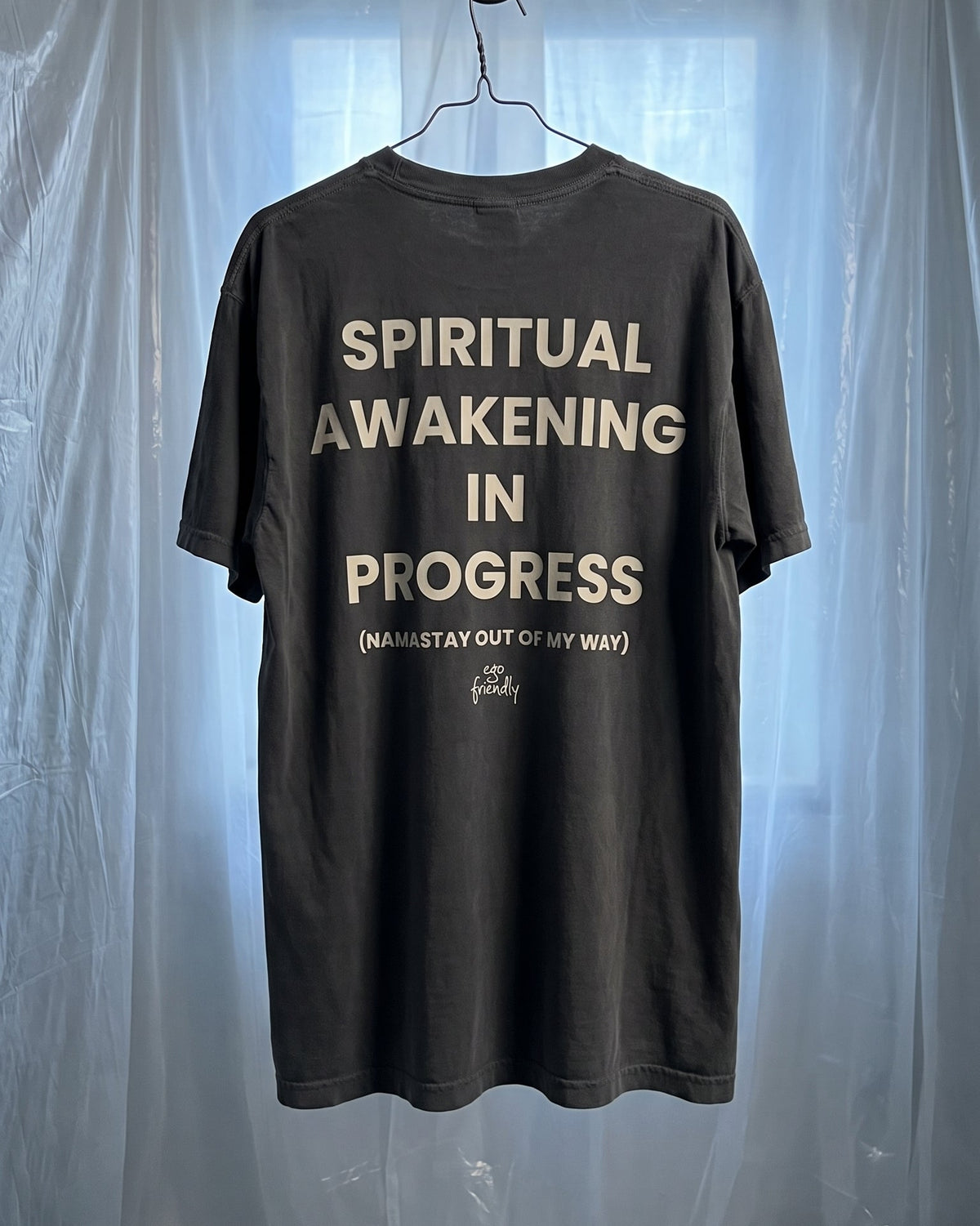 A dark charcoal gray t-shirt hanging on a metal hanger from a hook, featuring the text 'SPIRITUAL AWAKENING IN PROGRESS' in a light beige font on the back. The shirt is photographed against a backdrop of draped translucent plastic sheeting.