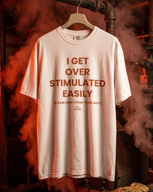 A white graphic t-shirt hanging on a wooden hanger against an industrial background with pipes, steam, and orange lighting. The shirt features the text 'I GET OVER STIMULATED EASILY' in brown, with smaller text below reading '(PLEASE DON'T PUSH YOUR LUCK) fate'.