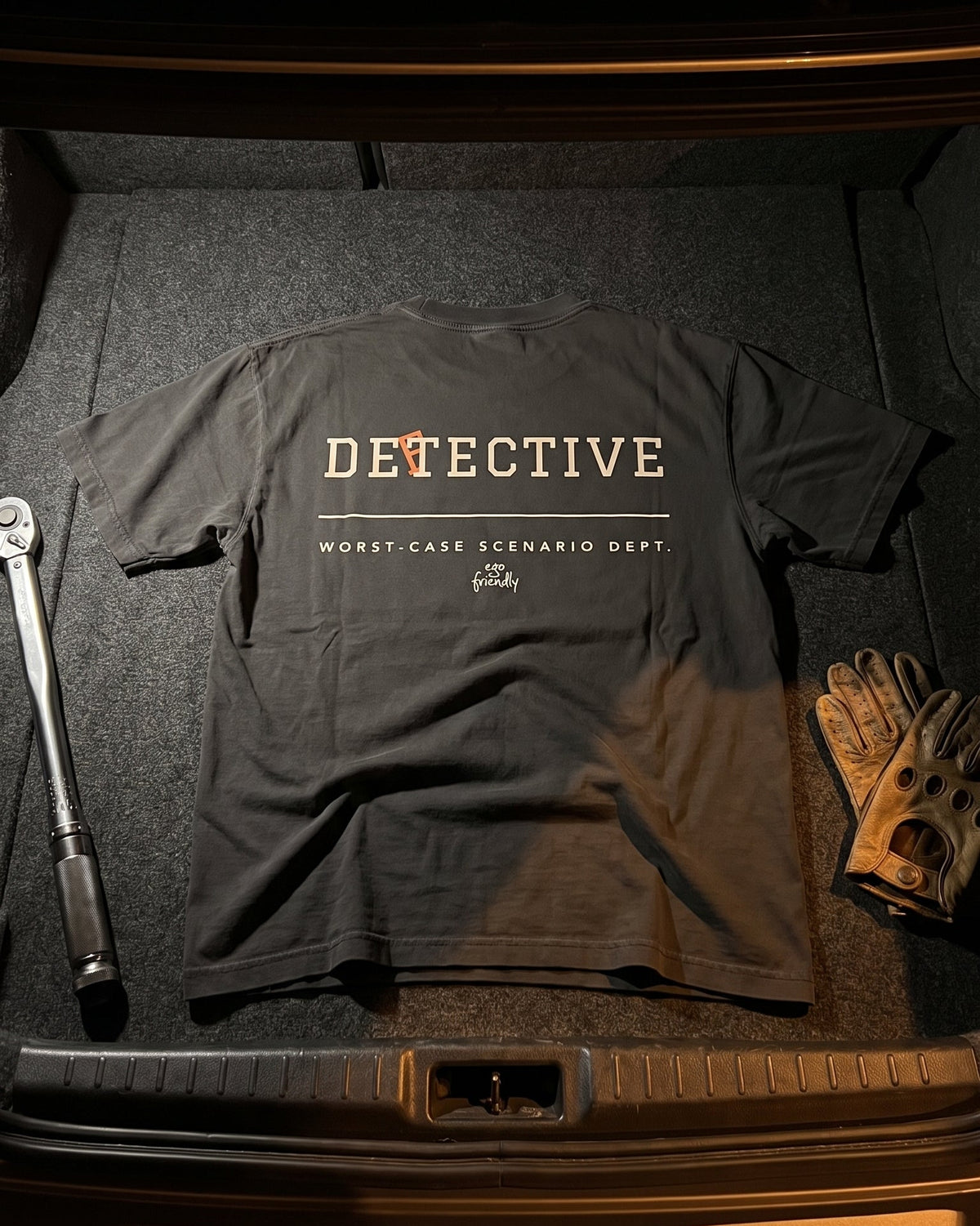 A black graphic t-shirt with 'DETECTIVE' printed in beige and orange across the chest, laid flat in a car trunk alongside a wrench and work gloves.