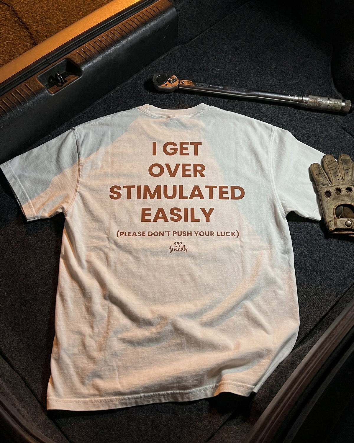 A white graphic t-shirt is laid flat on a dark textured surface, surrounded by a wrench, a work glove, and a leather tool case. The back of the shirt features brown text that reads 'I GET OVER STIMULATED EASILY (PLEASE DON'T PUSH YOUR LUCK)'.
